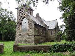 A photograph of a grey-bricked, many-windowed church with a slanted roof in a grassy field surrounded by green trees all under a bright sky