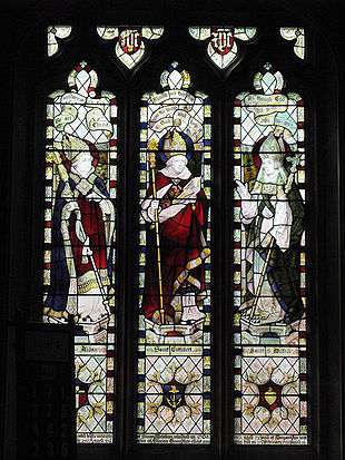 Three stained glass windows, each depciting a mitred and robed figure. All three are carrying staves.