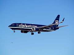 Left side view of an Alaska Airlines 737-800 painted in a primarily black livery, and says "alaskaair.com" on both sides. Aircraft is airborne.