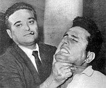 A black-and-white photograph showing a man wearing a jacket and tie with a short sigar in his mouth, on the left, holding up the chin of a man on the right, who appears to be in pain