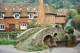 Narrow stone bridge over water. In the background are houses and trees.