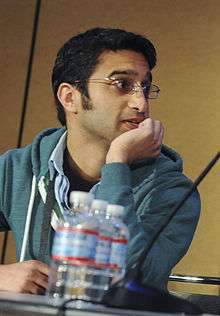 A man wearing a teal hoodie looking to the left. He is seated at a table behind a microphone and water bottles.