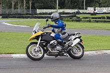 Black and yellow BMW R1200GS motorcycle, being ridden around a corner on a race track by a rider in a blue and grey suit
