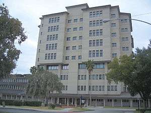 a ten story building with sable palms, a Spanish moss-drapped oak tree, hedges, and a court yard