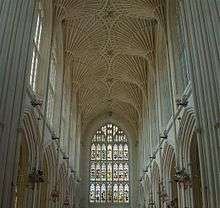 Interior of large building with a stained glass window at the far end. Above is a fan vault ceiling and on either side rows of arches.