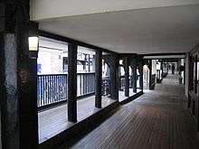An elevated covered walkway with the fronts of premises on the right and posts and railings overlooking the street on the left