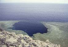 View of the coastal waters from the top of a hill, showing an approximately circular hole in the shallow coastal reef tangent to the deeper water offshore.