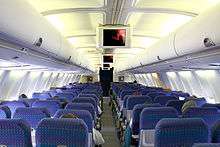Airliner cabin. Rows of seats arranged between a center aisle. There are overhead monitors.