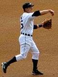 Baseball player in white uniform in a baseball throw followthrough position. His arm is forward with his hand down and his left hand is gloved. His left foot is planted and his right foot is in the air behind him.