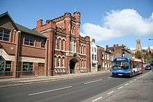 modern street scene, of broadgate with a bus heading toward us. But on the far side of the road is the astonishing mock-gothic frontage of the Drill Hall, in redbrick with stone details, including two turret towers, pierced with cruciform windows
