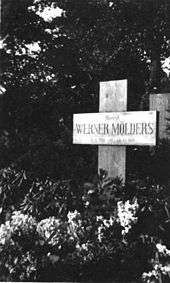 Black-and-white photograph of a wooden cross on a grave, bearing the inscription "Oberst Werner Mölders, 18. 3. 1913 – 22. 11. 1944." The name Werner Mölders is in large letters. Trees are seen in the background; the area in front of the cross is covered with low-growing plants bearing flowers.