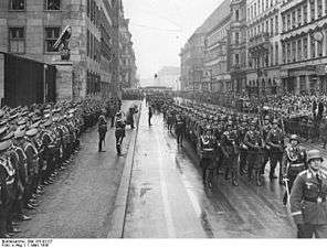 Soldiers with rifles and helmets march along a road past a reviewing party and guard of honor