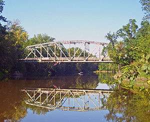 A pale metal bridge over brown water between two wooded areas