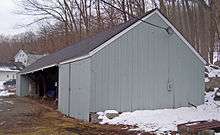 A long gray wooden building with a pointed black roof and mostly open front seen from near one end. There are patches of snow in the area.