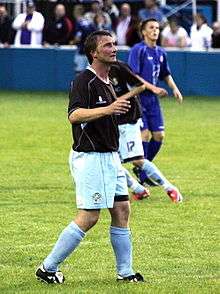 A man with dark hair who is wearing a black top, light blue shorts and light blue socks. He is standing on a grass field.