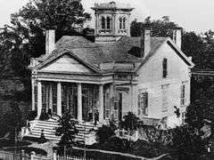 A white house with multiple chimneys and a tower. People are on the front porch and someone is arriving at the home and about to embark up the staircase. The front porch has a roof over a set of four columns. Trees are visible on the lawn.
