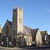 A pale stone church on a corner site, with a dominant square tower with spirelets. To the left of this is the entrance, below a four-light lancet window and three sexfoils. To the right is the main body of the church, with two side chapels and lancet windows.