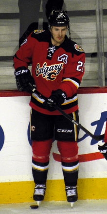 A hockey player in full uniform. The Jersey and legs are red with black, white and yellow trim, while the pants and helmet are black. The logo says "Calgary" in script and includes a small stylized "C".
