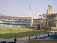 Back view of the entrance and a stand of a cricket stadium. Some people can be seen near the seats.