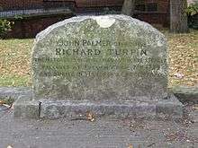 A stone gravestone with a curved top, with several lines of inscription. Trees, grass, and a wall are visible in the distance.