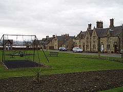 A village green with a swing set in the middle and a row of houses in the background