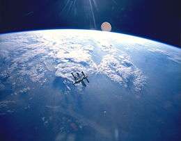 A cluster of modules and feathery solar arrays floats in the middle distance before an image of the Earth and the blackness of space above its horizon. Sunrays project from the top centre of the image.