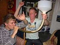 One person holds a clear pipe to their mouth, while a second holds the other end of the pipe with a large white plastic funnel attached. The pipe is part-filled with a translucent yellow-brown liquid, with the remainder of the pipe and the bottom of the funnel filled with a foamy head.