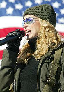 A blonde woman wearing a woolly hat and glasses singing into a microphone in front of the American flag