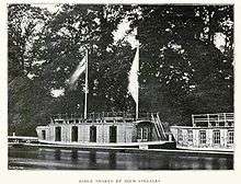 Two barges by the side of a river, one not fully in shot, in front of trees. The main barge has two large flags flying from the cabin roof, four large openings and one small one on the cabin's river side, and a window on the side of the cabin perpendicular to the river alongside a staircase to the roof; there are railings around the top of the roof. The other barge has five visible windows and roof railings.