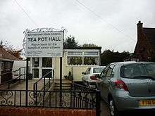 A flat-roofed temporary building,neatly finished in cream. A ramp for 'disabled access' leads to the door, and two cars are parked on the right. Prominent on the left a sign decorated with 'wrought' iron scrolls reads Bottesford town council, TEA POT HALL, pop-in room for the benefit of senior citizens