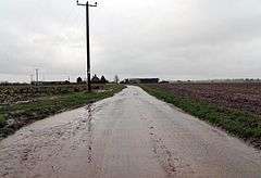A completely flat landscape. A wet, muddy road stretches in the distance, dead straight, flanked by empty, wet fields. A wooden electricity pole is a sudden vertical element. The wires follow the road down to some farm buildings in the middle distance