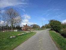 A sunny day in a green landscape, under a blue sky. A country lane meanders from the middle foreground to traffic lights in the right midground. On the right is a grass verge and a hedge. On the left, a grass verge edged with large boulders. Beyond that a wooden rail fence marks the edge of a smaller road, and beyond that a building with a ridged pantile roof and large ornate bargeboards. This is 'station house' the last remnant of the railway hereabouts.