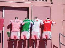 Four cycling jerseys – pink, green, white, and red, from left to right – are arranged on mannequin torsos in front of a pink wall.