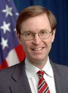 A Caucasian male posing for a professional photo wearing a gray suit with a white dress shirt and red, white and blue striped tie. An American flag is seen in the background.