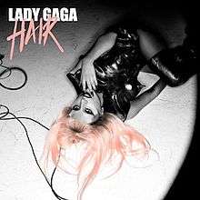 Greyscale upturned image of Lady Gaga lying on the ground, wearing a black leather dress. Her left hand is near her mouth and she looks up to the camera. On top-right the words "Lady Gaga" and "Hair" are written. Her flowing hair and the word "Hair" are colored pink.