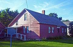 A 1 1⁄2-story wooden house, painted red, lit by the sun from the left with trees and part of a white house behind it.
