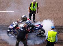 A silver and blue sport motorcycle with racing numbers and covered sponsor logos. The rider has a reflective helmet and wears brightly colored leathers also covered with logos. His hands on the clutch and brake levers while the front wheel does not turn and the rear wheel spins and emits a cloud of white smoke. Three officials stand close by and observe.
