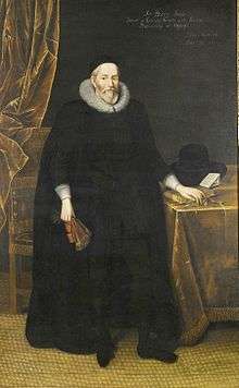 A tall elderly man with a beard, wearing long black robes and a large white ruff. He is standing with a fan in his right hand and with his left hand resting on books on a table.