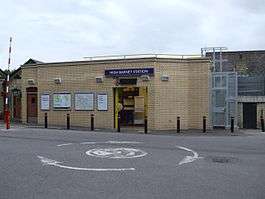 A beige-bricked building with a rectangular, blue sign reading "HIGH BARNET STATION" in white letters all under a blue sky with white clouds