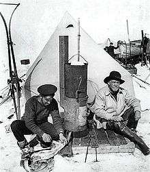 Two men sitting in front of a pointed tent; left-hand man in flat naval cap is skinning a penguin, right-hand man wears a wide-brimmed hat. Between them is a tall stove. Other equipment is visible in the background
