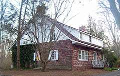 A house built of red stone house with a curved, sloping roof and white wooden top, seen from the side. It has brick chimneys at either end and some ivy on the visible side.