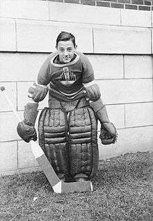 A teenage Plante assumes the traditional goaltender stance, slightly crouched with legs together, wearing goaltender pads on his leg, his team sweater, and holding a goaltender stick in his right hand with the blade of the stick in front of his feet