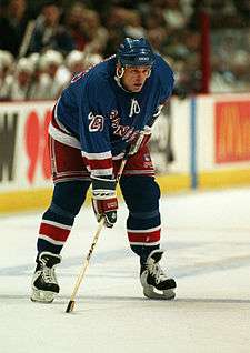 A Caucasian hockey player. He wears a dark blue jersey and helmet. He is bent over at the waist leaning on his stick.