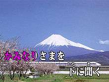 Over a background image of Mount Fuji and a Shinkansen train two lines of Japanese text. The first half of the first line is colored red-violet, the rest white. The text reads "かみなりさまを 下 に聞く".