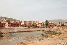 A river runs across the image from left to right with a town in the background, behind a concrete flood defence. The foreground shows a stony, sparsely vegetated river bank.