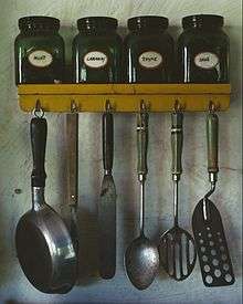 Various kitchen utensils. At top: a spice rack with jars of mint, caraway, thyme, and sage. Lower: hanging from hooks; a small pan, a meat fork, an icing spatula, a whole spoon, a slotted spoon, and a perforated spatula.