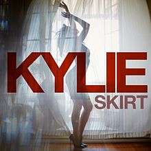 Minogue standing in heels behind a curtain, with the words 'Kylie' and 'Skirt' superimposed in a red font.