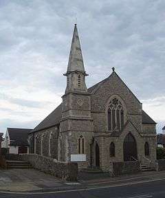A pale stone building with a tiled roof sloping further on the left than on the right. A tower with a two-light lancet window, diamond-shaped decoration and a short spire stands in the near corner. A porch, with two doors and two windows in a lancet style, stands in front of the main body of the church, dominated by a four-light lancet.