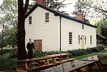 A white house in three-quarter perspective. In the foreground are several picnic benches. To the left is a tall tree with a man standing beside it. To the right, partly obsured by the house, is an old well.
