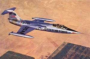 Jet fighter in metallic scheme with T-tail and short wings flying above desert and black constructions